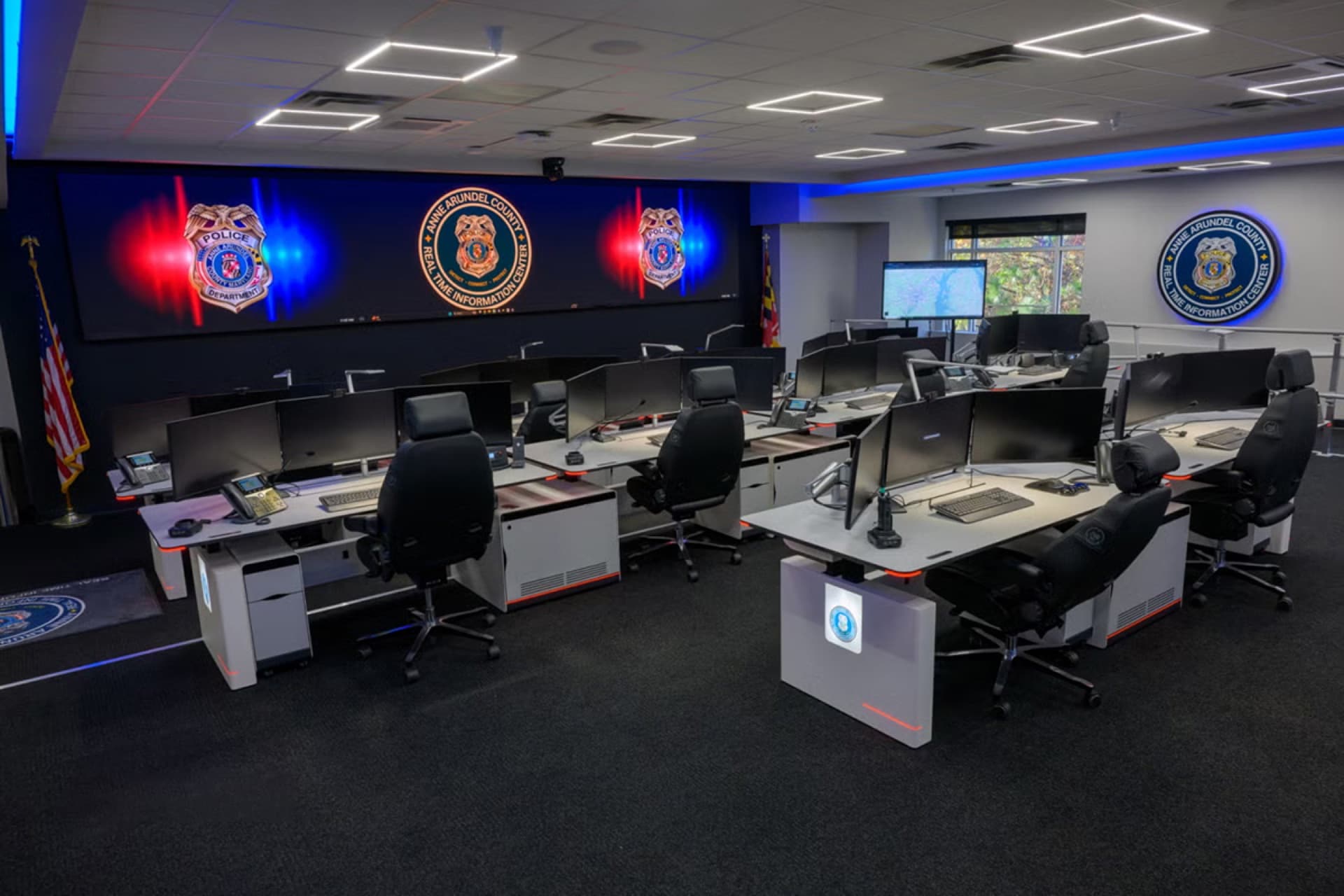 Anne Arundel County Police real-time crime center with white sit-stand dispatch consoles and a video wall of agency badges